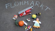 Verschiedenes Spielzeug liegt auf dem Boden, darüber steht mit Kreide Flohmarkt geschrieben. © NDR Foto: Julia Henke