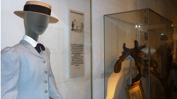 Eine Puppe mit einem Strohhut und einem Blazer steht vor einer Vitrine, in der ein Pferdesattel zu sehen ist. © Landkreis Verden 