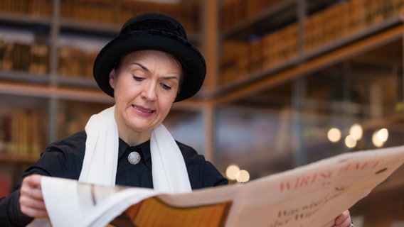 Eine Frau mit einem schwarzen Zylinder auf dem Kopf liest Zeitung und schaut dabei ernst. © Landkreis Verden 