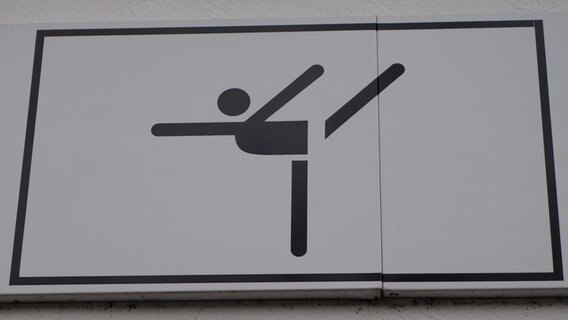 Auf einem Schild ist ein stilisiertes Männchen zu sehen, das ein Bein nach hinten in die Luft streckt. © NDR Foto: Andrea Heußinger