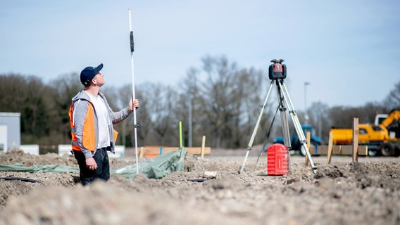 Henry Heidrich, Auszubildender in einem Bauunternehmen, steht auf einer Baustelle und misst die Tiefe eines Grabens für das Fundament mit einem Rotationslaser. © picture alliance/dpa Foto: Hauke-Christian Dittrich