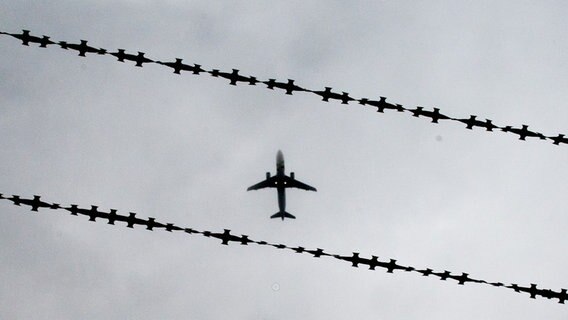 Ein fliegendes Flugzeug ist von unten zu sehen. Davor Stacheldraht. © Julian Stratenschulte/dpa Foto: Julian Stratenschulte
