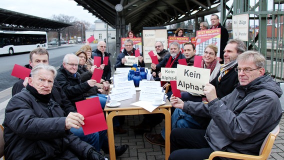 Menschen sitzen an einer Haltestelle um einen Tisch herum und zeigen rote Karten. © Hansestadt Lüneburg Foto: Hansestadt Lüneburg