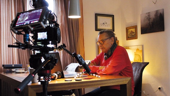 Der Schauspieler sitzt an einem Tisch, neben ihm steht eine Filmkamera auf einem Stativ. © Salzgeber & Co. Medien GmbH 