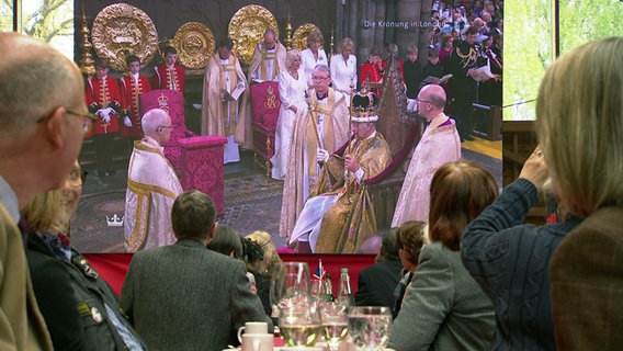 Menschen schauen bei einer Veranstaltung der Hannoversch-Britischen Gesellschaft gemeinsam die Krönung von König Charles auf einer Leinwand. © NDR 