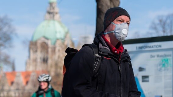 Ein Mann mit Atemschutzmaske steht in Hannover in der Sonne, im Hintergrund ist das Neue Rathaus zu sehen. © dpa - Bildfunk Foto: Julian Stratenschulte
