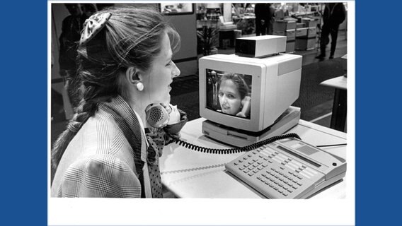 Ein schwarz/weiß Aufnahme zeigt eine Frau an einem alten Computer und einem Videobild von ihr auf einem Bildschirm. © Deutsche Messe AG 