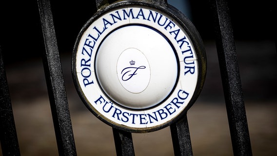Das Logo der Porzellanmanufaktur Fürstenberg ist an einem Zaun vor dem Unternehmen zu sehen. © Moritz Frankenberg/dpa Foto: Moritz Frankenberg