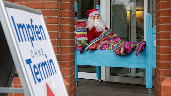 Eine Weihnachtsmann-Puppe sitzt in einem Bett als Werbung vor einem Bettengeschäft, im Vordergrund ein Hinweisschildes "Impfen ohne Termin" eines Impfzentrums. © dpa-Bildfunk Foto: Julian Stratenschulte/dpa