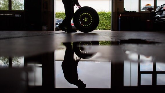Ein Auszubildender zum KfZ-Mechatroniker rollt einen Reifen durch eine Werkstatt. © dpa Foto: Julian Stratenschulte
