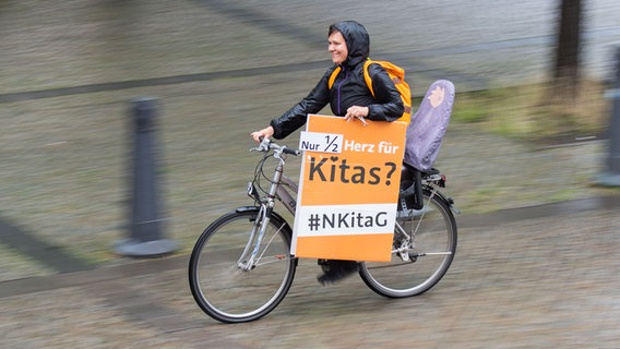 Eine Frau radelt mit einem Plakat mit der Aufschrift "Nur 1/2 Herz für Kitas" vor dem niedersächsischen Landtag entlang. © dpa-Bildfunk Foto: Julian Stratenschulte