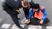 Polizisten versuchen festgeklebte Hände von Aktivisten der Gruppierung "Letzte Generation" am Deisterkreisel in Hannover mit Speiseöl von der Straße abzulösen. © picture alliance/dpa Foto: Julian Stratenschulte