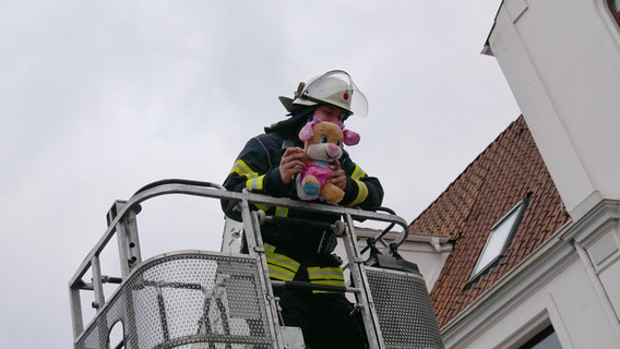Eine Einsatzkraft der Feuerwehr steht auf einer Drehleiter und rettet ein Kuscheltier aus einer Dachrinne. © Feuerwehr Celle/dpa 