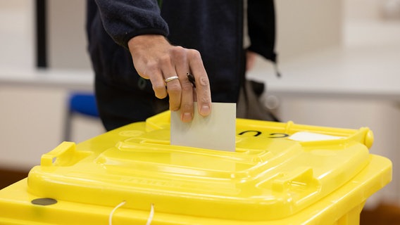 Ein Wähler wirft ihren Stimmzettel in eine Tonne mit Schlitz im gelben Deckel (Wahlurne). © Friso Gentsch/dpa Foto: Friso Gentsch/dpa
