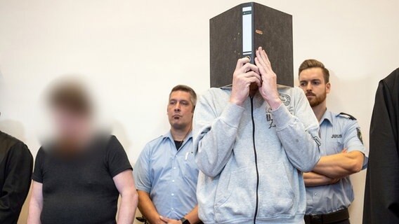Der Angeklagte Andreas V. steht im Gerichtssaal und hält sich einen Aktenordner vor das Gesicht, daneben steht der Angeklagte Mario S. © dpa - Bildfunk Foto: Bernd Thissen