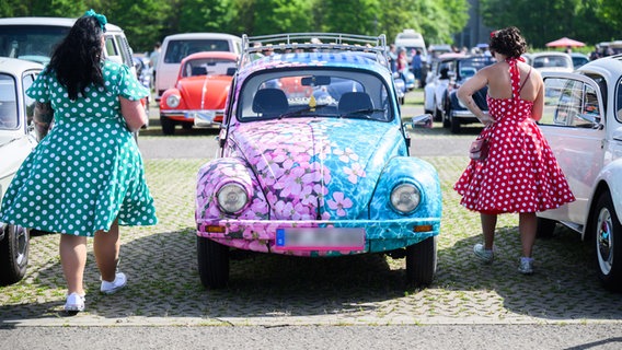 Zwei Frauen in Kleidern laufen neben einem Volkswagen Käfer im Blümchenoptik beim MaiKäfertreffen auf dem Messegelände. © Julian Stratenschulte/dpa 