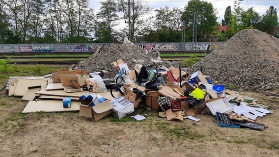 Ein zunächst Unbekannter hat auf einem Bahngelände illegal Müll abgeladen. Der Müllberg liegt vor zwei Erdhaufen. Im Hintergrund sind Gleise und eine bemalte Lärmschutzwand zu sehen. © Bundespolizei Hannover Foto: Bundespolizei Hannover