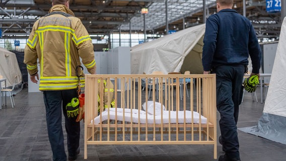 Feuerwehrleute tragen ein Kinderbett in ein sogenanntes Dorf, ein abgetrennter Bereich mit vier Zelten, Spinden und Kühlschränken für die Geflüchteten. © dpa Foto: Lino Mirgeler
