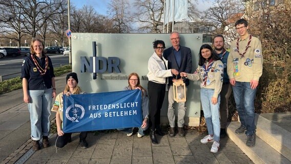 Vor dem NDR-Schild stehen Pfadfinder und halten ein Transparent mit der Aufschrift "Friedenslicht aus Betlehem", ein Mädchen übergibt Funkhausdirektorin Andrea Lütke ein Friedenslicht. © NDR Foto: Marco Heuer