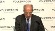 Hans Dieter Pötsch sitzt vor einem Aufsteller auf dem "Volkswagen" steht. © NDR 