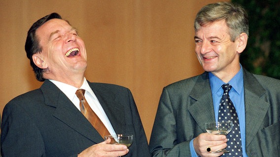 Lachend und mit einem Sektglas in der Hand zeigen sich Gerhard Schröder und Joschka Fischer nach der Unterzeichnung des rot-grünen Koalitionsvertrages in der NRW-Landesvertretung in Bonn 1998. © picture alliance / dpa Foto: Michael Jung