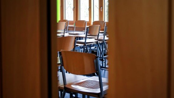 Durch den Spalt einer offenstehenden Tür ist ein leeres Klassenzimmer mit hochgestellten Stühlen zu sehen. © dpa - Bildfunk Foto: Henning Kaiser