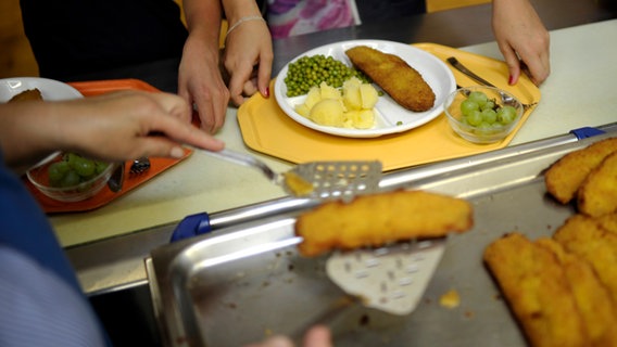 Eine Küchenhilfe legt ein Stück panierten Fisch auf einen Teller eines Schulkindes in einer Schulküche. © dpa-Bildfunk Foto: Alexander Körner