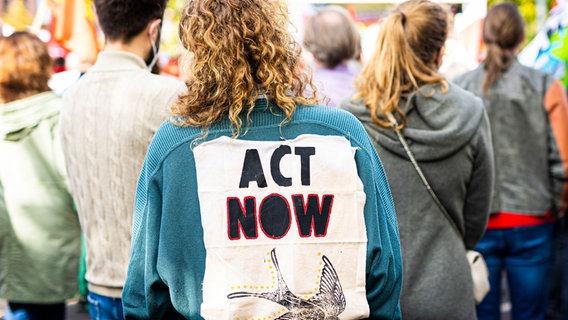 "Act now" steht auf dem Zettelm den eine Demonstrierende auf dem Rücken trägt. © Moritz Frankenberg/dpa Foto: Moritz Frankenberg/dpa