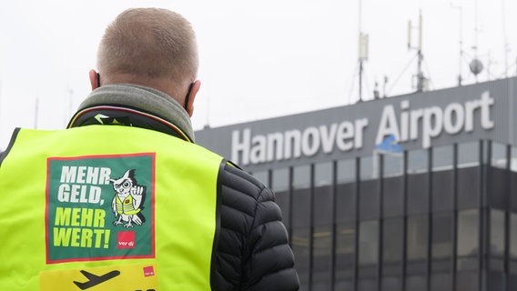 Ein Flughafen-Mitarbeiter steht mit einer ver.di-Warnweste vor dem Flughafen in Hannover. © picture alliance Foto: Julian Stratenschulte