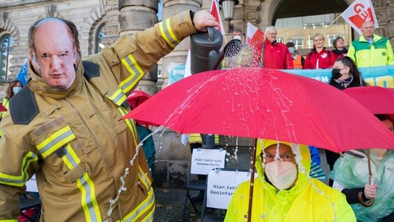 Ein Feuerwehrmann mit einer Maske vom Finanzminister Niedersachsens Hilbers gießt Wasser über einen Kollegen vor dem Finanzministerium. © picture alliance Foto: picture alliance/dpa | Julian Stratenschulte