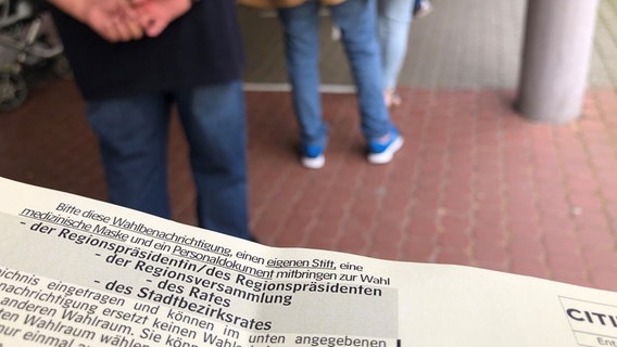 Ein Stück einer Wahlbenachrichtigung ist vor einer Menschenschlange zu sehen. © NDR Foto: Angelika Henkel