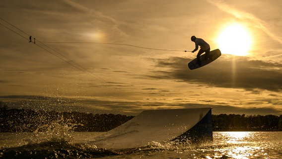 Vor dem Sonnenuntergang springt ein Wakeboarder über eine Rampe. © dpa - Bildfunk Foto: Christophe Gateau