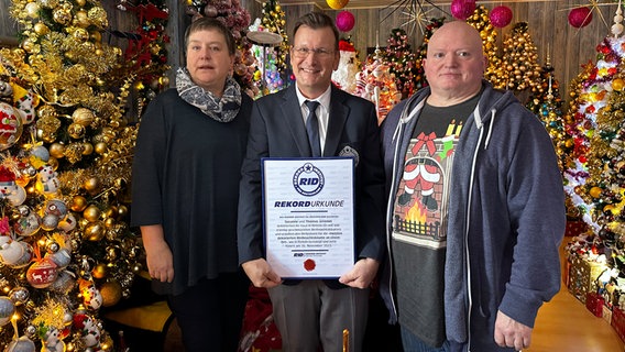 Susanne und Thomas Jeromin stehen mit einem  Prüfer des Rekord-Instituts für Deutschland (Mitte) in ihrem Haus inmitten von Weihnachtsbäumen. © NDR Foto: Larissa Mass