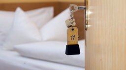 Ein Schlüssel mit einem Anhänger mit der Nummer 11 steckt in einer geöffneten Hotelzimmertür, im Hintergrund ist ein Bett zu sehen.