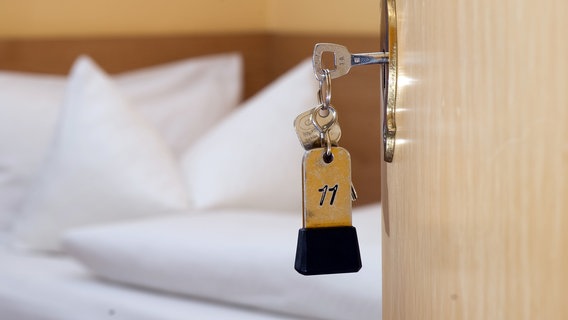 Ein Zimmerschlüssel hängt in einem Gästehaus vor einem Bett im Türschloss. © dpa-Bildfunk Foto: Swen Pförtner