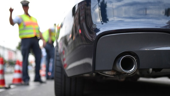 Im Zentrum des Bildes steht der Auspuff eines Autos in der Unschärfe vor dem Auto steht ein Polizist. © picture alliance / dpa Foto: Uwe Anspach