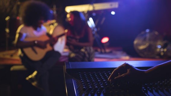 Eine Hand betätigt einen Regler an einem Mischpult. Im Hintergrund spielt ein Gitarrist. © dpa - picture alliance 