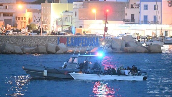 Ein Polizeiboot und ein Boot mit geflüchtete Menschen fahren in den Hafen von Lampedusa ein. Im Hintergrund ist die Silhouette der Stadt zu sehen. © picture alliance / ANSA | CIRO FUSCO Foto: CIRO FUSCO