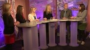 Eine Moderatorin steht mit 5 jungen Menschen an Hochtischen in einem Fernsehstudio. Im Hintergrund sind Wände aufgestellt, auf den Schlagzeilen aus Zeitungen aufgeklebt sind. © NDR 