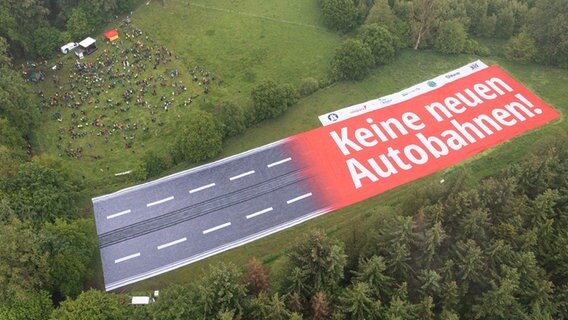 Aus der Luft ist ein Banner mit der Aufschrift "Keine neuen Autobahnen!" auf einer Landfläche zu sehen, daneben viele Menschen. © dpa Foto: Mohssen Assanimoghaddam