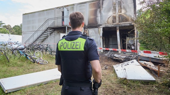 Ein Polizist steht vor einer abgebrannten Unterkunft für Geflüchtete. Ein Kollege wurde schwer verletzt. © picture alliance/dpa Foto: Georg Wendt