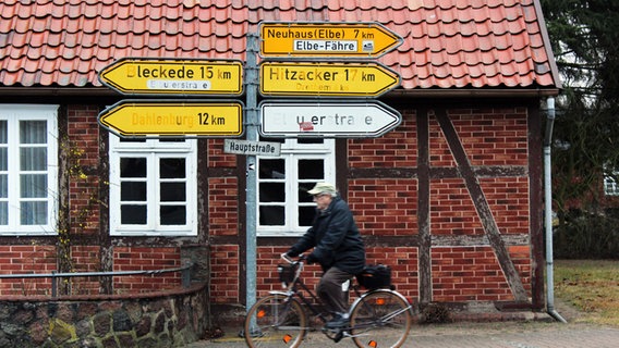 Verkehrsschilder unter anderem mit der Aufschrift: Neuhaus (Elbe) 7 km. Ein Fahrradfahrer fährt an den Schildern vorbei. Verkehrsschilder unter anderem mit der Aufschrift: Neuhaus (Elbe) 7 km. Ein Fahrradfahrer fährt an den Schildern vorbei. © NDR Foto: Ann-Kristin Mennen