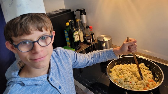 Ein Junge mit Kochmütze rührt mit einem Kochlöffel in einer Pfanne mit Rührei und schaut in die Kamera. © NDR Foto: Helene Buscholz