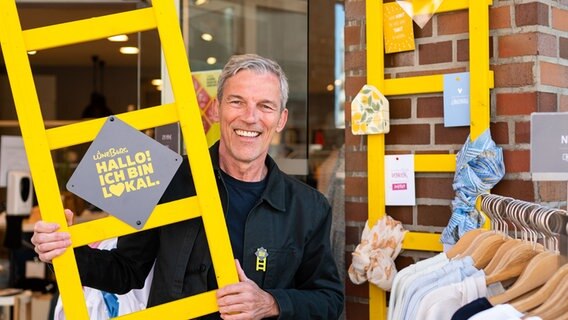 Ralf Elfers, Ideengeber und Ladenbesitzer, steht mit einer gelben Leiter mit einem Schild "Hallo! Ich bin lokal." vor seinem Geschäft in der Innenstadt Lüneburg. © dpa-Bildfunk Foto: Philipp Schulze