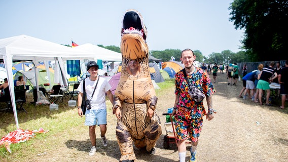 Drei Personen gegen nebeneinander über ein Festival-Gelände. Eine Person trägt ein Dinosaurierkostüm. © Hauke-Christian Dittrich/dpa Foto: Hauke-Christian Dittrich