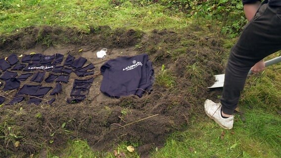 Stofffetzen und ein Pullover mit Aufschrift der Leuphana Universität werden in einer Erdgrube vergraben. © NDR 