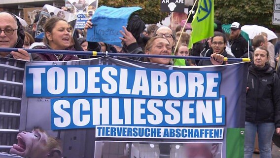 Eine Gruppe Menschen hat sich am Abend zu einer Mahnwache versammelt. Auf einem Schild steht "Todeslabore schließen!" © NDR 