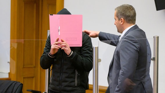Lüneburg: Der Angeklagte, der sein Gesicht hinter einer Aktenmappe verbirgt, wird  am 09.11.2022 von Moritz Klay, seinem Anwalt, in den Saal des Landgerichtes geführt. © dpa-Bildfunk Foto: Philipp Schulze