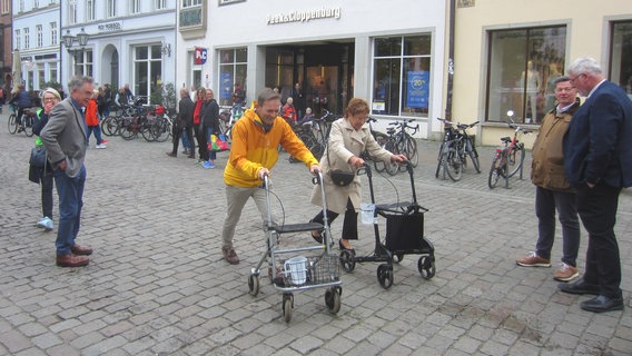 Zwei Personen fahren mit einem Rollator über Kopfsteinpflaster und versuchen dabei, möglichst wenig Wasser aus Gefäßen, die am Rollator befestigt sind, zu verschütten. © FUSS e.V. Lüneburg Foto: J. Korn