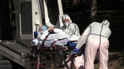 Zwei Männer in Schutzanzügen tragen einen Mann auf einer Liege in einen Krankenwagen. 
